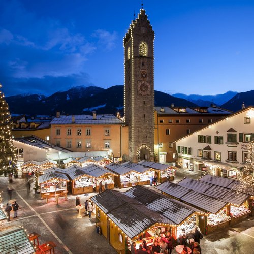 Das Skigebiet Ladurns und andere Winter-Highlights Beleuchteter Weihnachtsmarkt mit Ständen und großem Tannenbaum bei Abenddämmerung