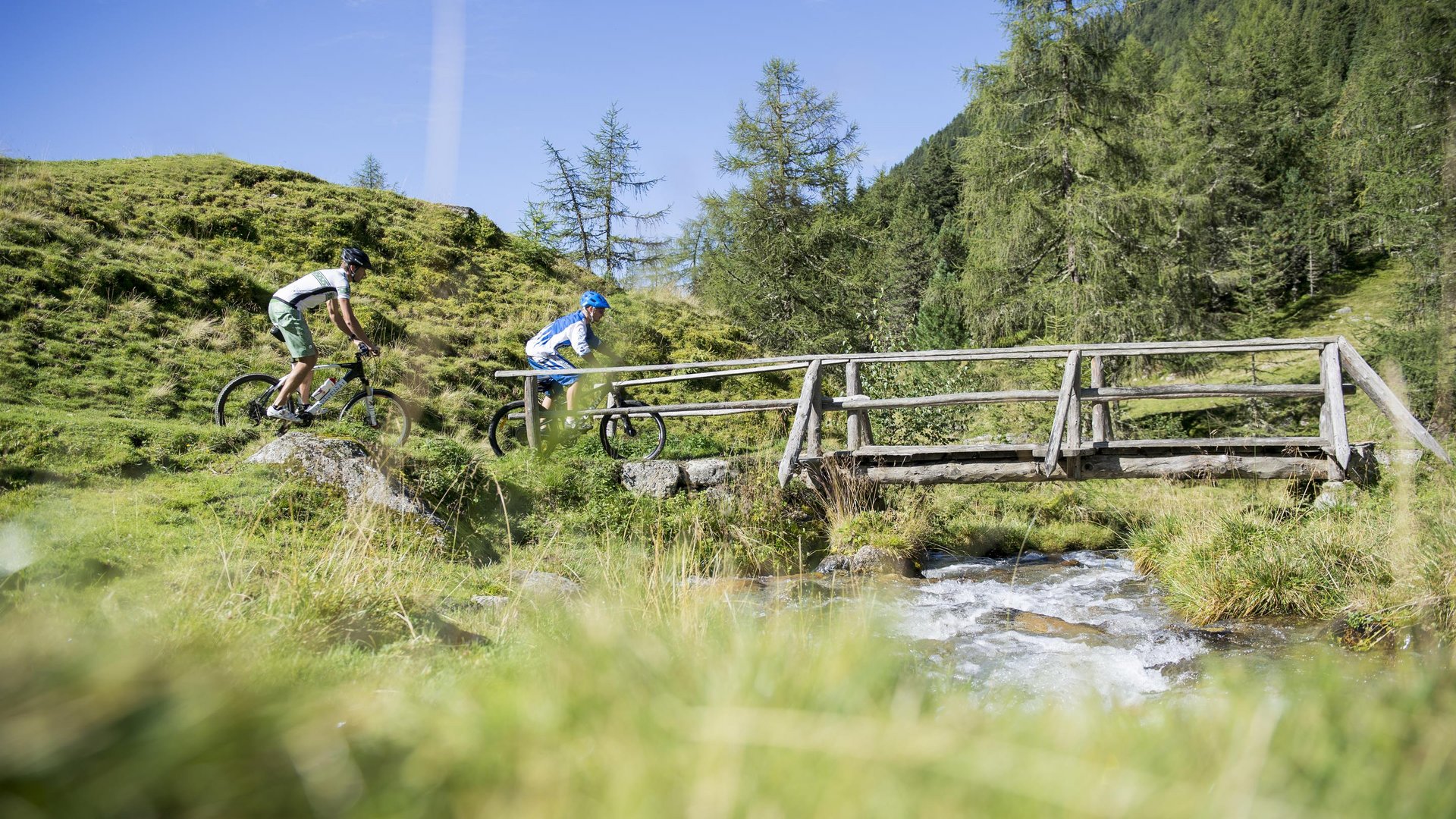 Wandern in Sterzing und Umgebung Zwei Radfahrer überqueren eine Holzbrücke über einen Bach in den Bergen