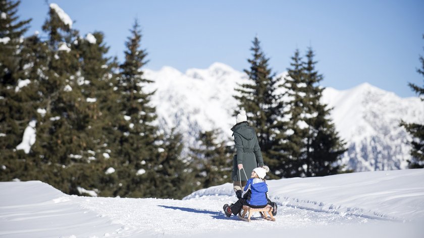 Wintererlebnisse by Gudrun Frau zieht Kind auf Schlitten durch verschneite Winterlandschaft mit Tannen