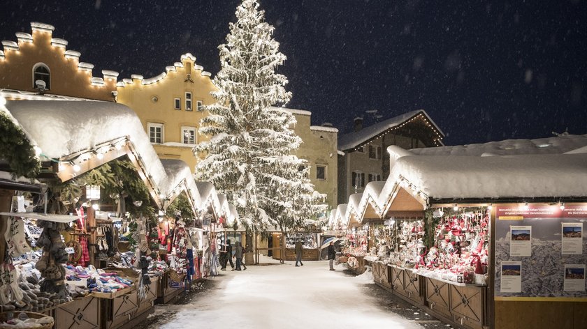 Die „Sterzinger Glockenweihnacht“ Verschneiter Weihnachtsmarkt mit beleuchtetem Tannenbaum und Hütten bei Nacht