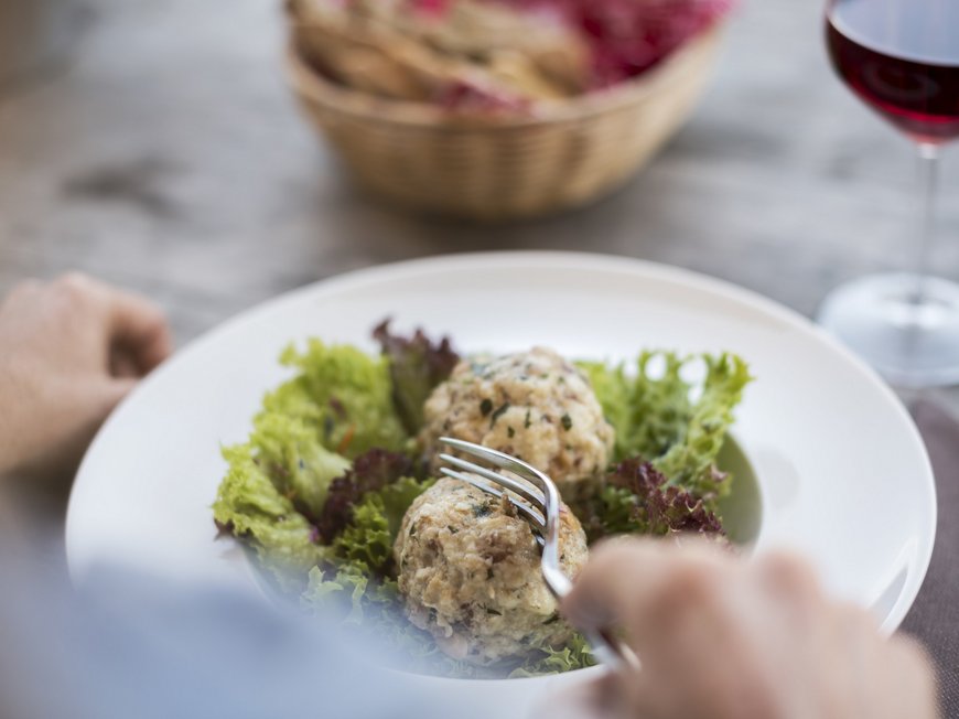 Wandern in Sterzing und Umgebung Person isst zwei Knödel auf Salat mit Glas Rotwein und Brotkorb im Hintergrund