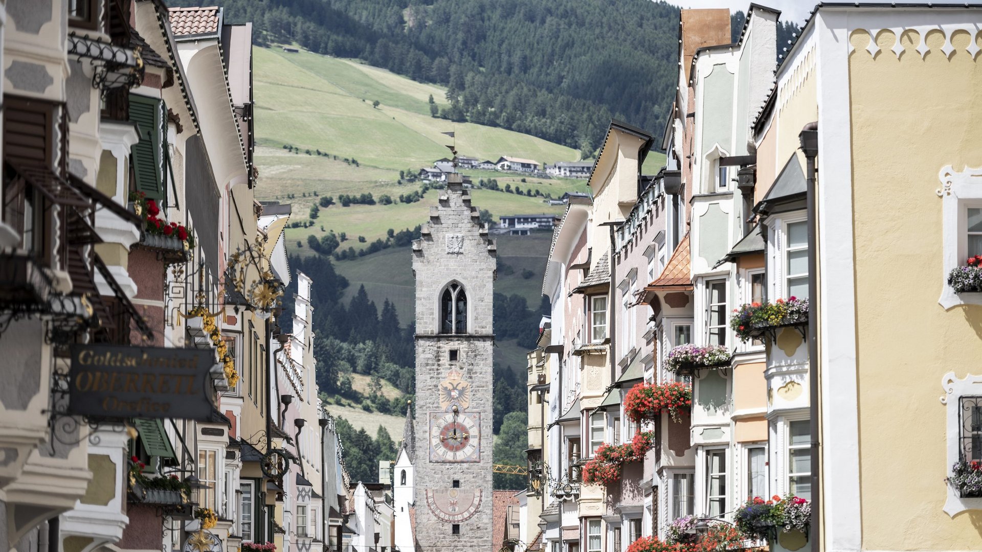 Hotel in Val di Fleres: gli eventi Case colorate con fiori su una strada davanti a una torre dell'orologio e montagne
