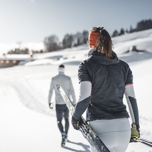 Das Skigebiet Ladurns und andere Winter-Highlights Frau trägt Skier auf verschneiter Loipe, Mann geht voraus vor Bergdorf