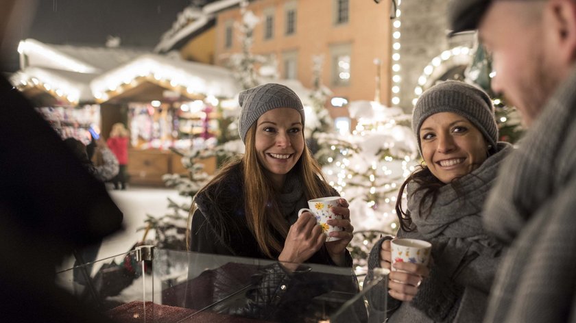Gudrun stößt an: mit Glühwein Lächelnde Freunde trinken Heißgetränke auf einem winterlichen Weihnachtsmarkt