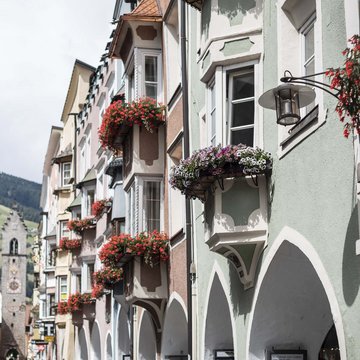 Gudruns Blog: alles über Sterzing, übers Wandern, über Tirol Bunte Häuser mit Blumen in Balkonen und Bogen auf einer Straße mit Bergkulisse