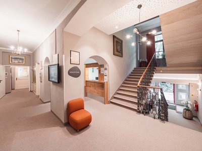 Hotel a Colle Isarco: benvenuti da Gudrun! Ingresso luminoso dell'hotel con reception, scala e poltrona arancione.